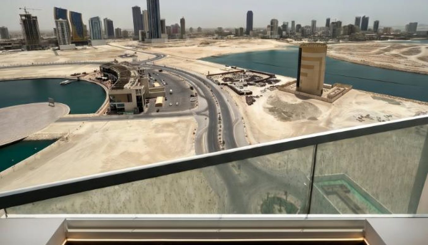 Cityscape with modern buildings, roads, and water bodies viewed from a high balcony with a glass railing and stone flooring in the foreground.