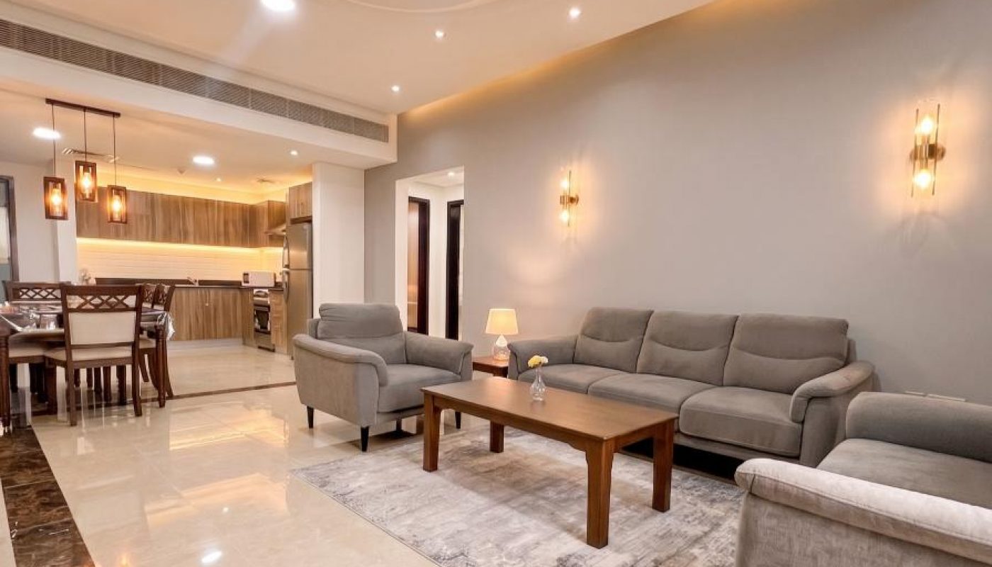 Modern living room with gray sofas, a wooden coffee table, wall sconces, and recessed ceiling lights, leading to an open kitchen and dining area with marble-tiled flooring.