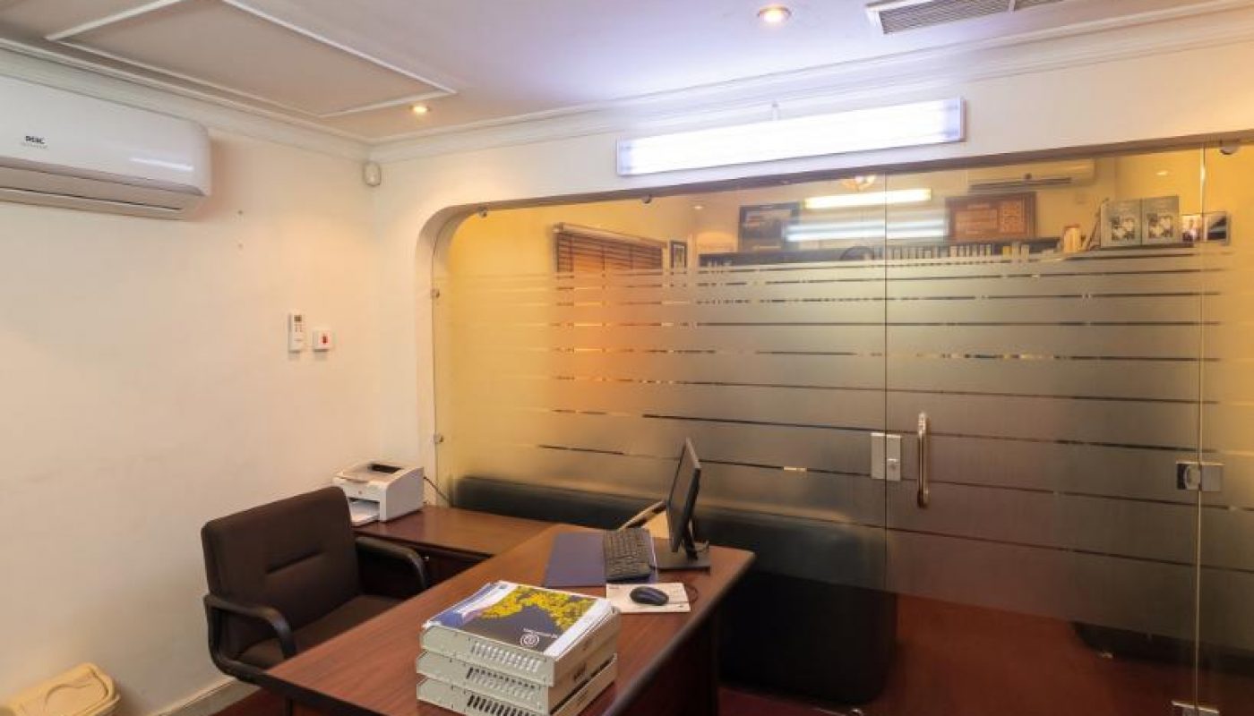 Office with a desk, chair, computer monitor, printer, and stacked files; glass partition with frosted stripes separates the space.