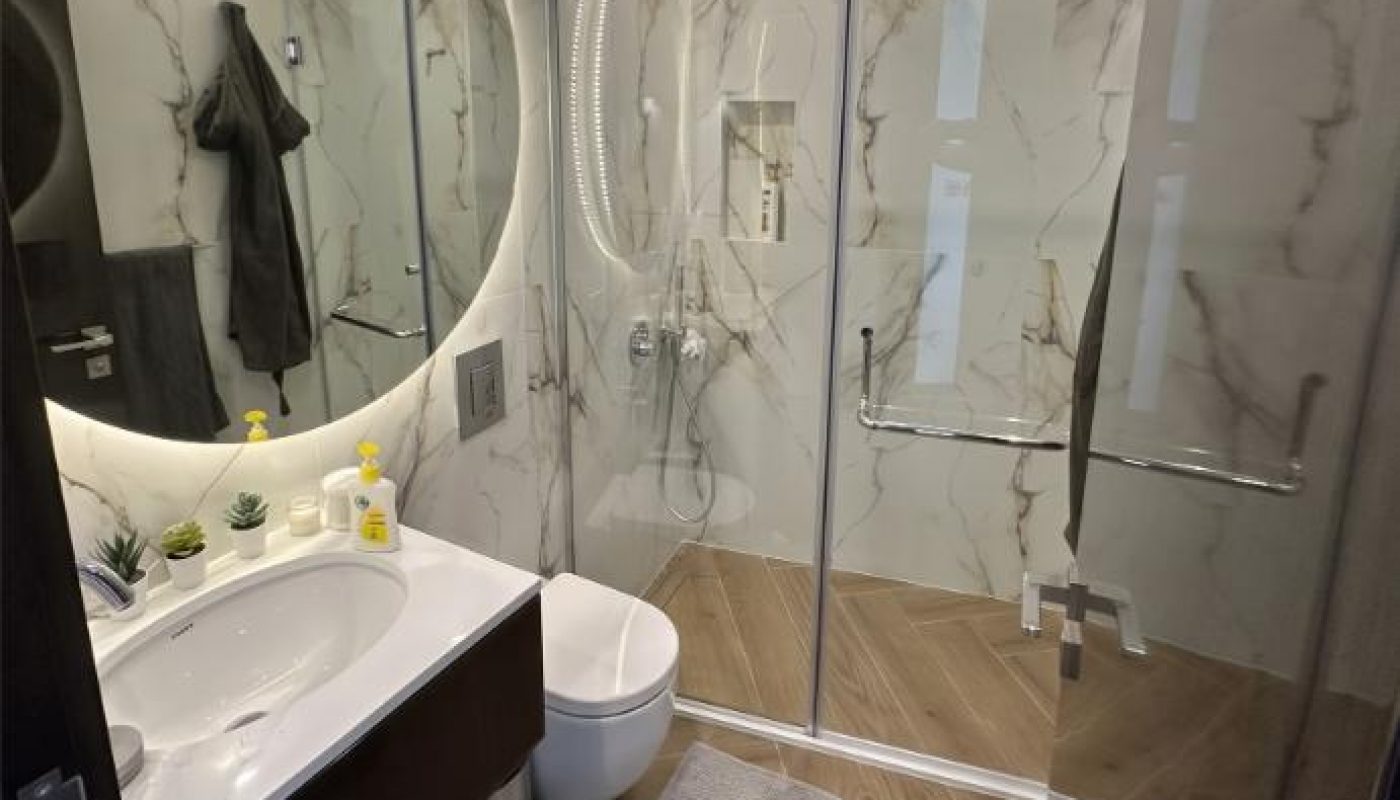 Modern bathroom with a round mirror, sink, toilet, glass shower enclosure, towel rack, and gray floor mat on wood-patterned tiles.