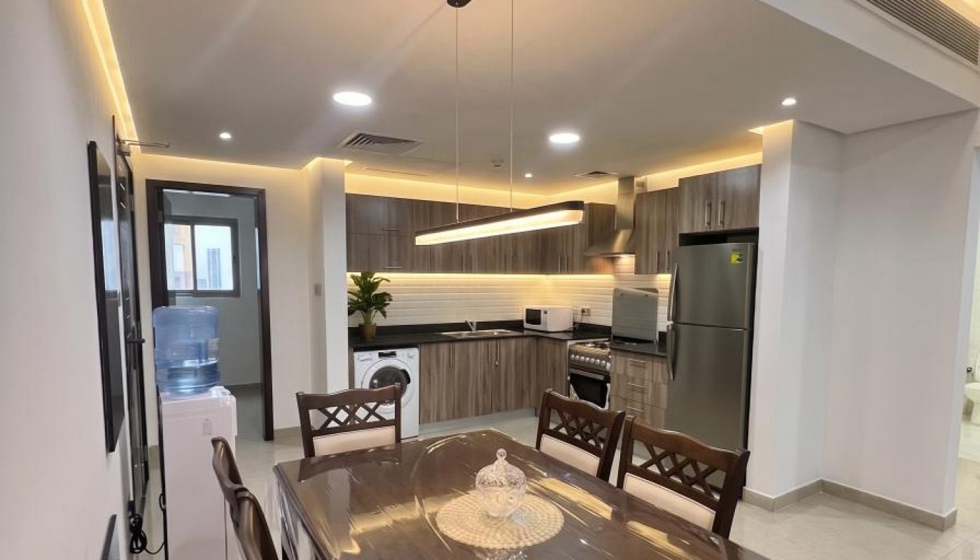 Modern dining area with a wooden table and six chairs, adjacent to a kitchen with wood cabinets, stainless steel appliances, and bright overhead lighting.