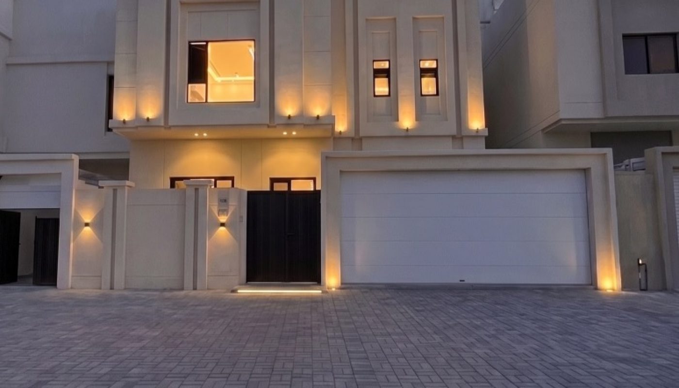 Modern two-story house with exterior lights, large garage door, and paved driveway, photographed at dusk under a partly cloudy sky.