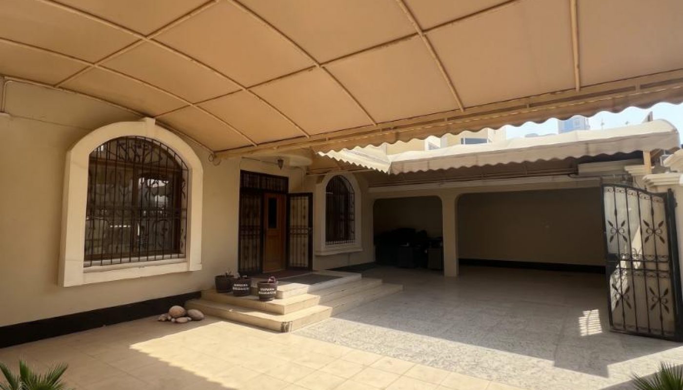 Covered house entrance with tiled floor, metal gate, wide steps, and small potted plants near the doorway under a tan canopy.