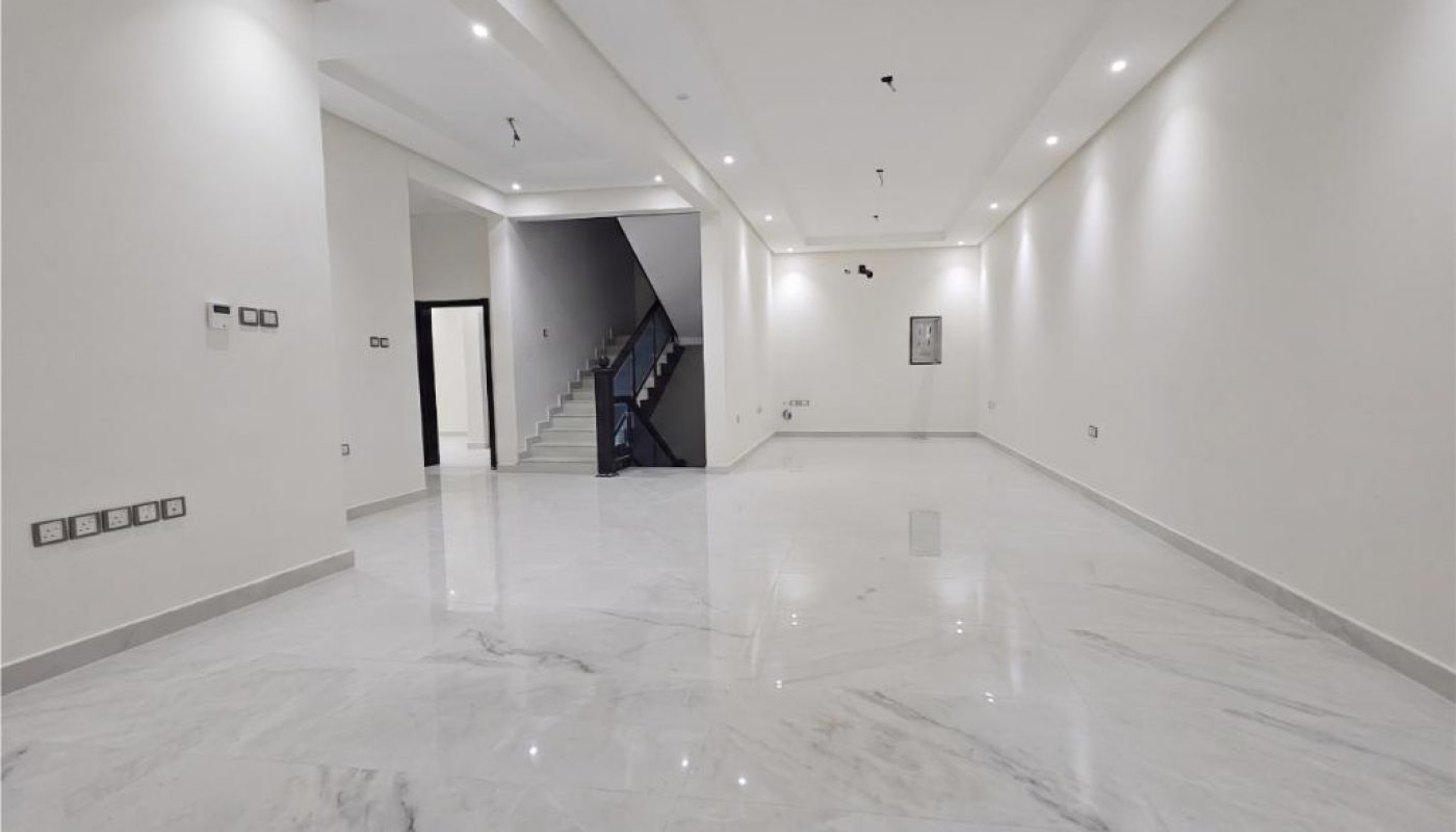 Spacious, empty room with white walls, recessed ceiling lights, marble floors, and a staircase leading to an upper floor in the background.