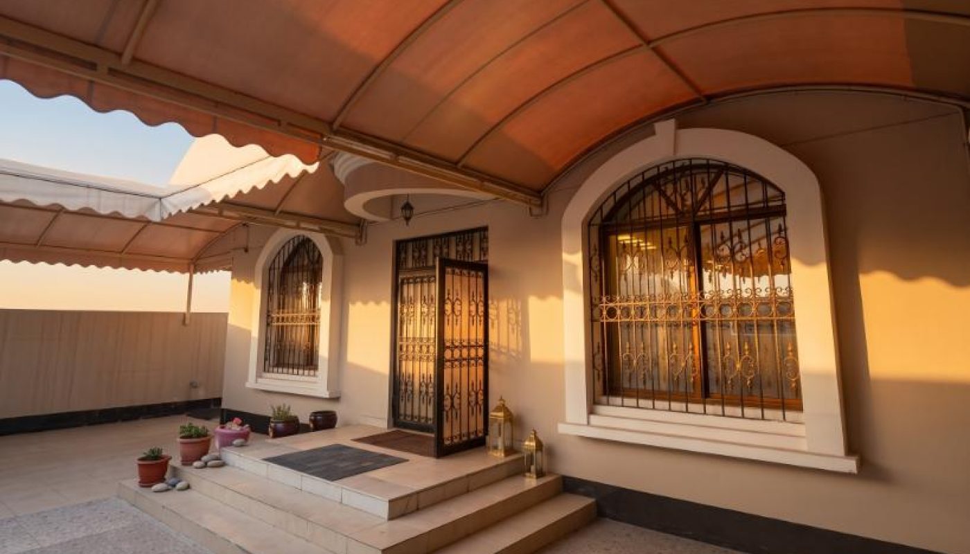 A covered outdoor patio with tiled floor, steps leading to a door with metal bars, arched windows, and several potted plants on the left.