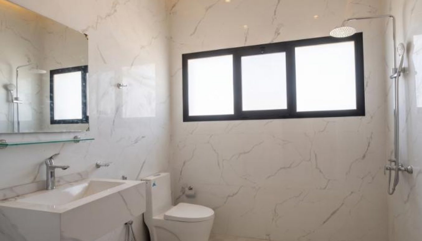 Modern bathroom with marble walls and floor, a floating sink, toilet, shower area, large mirror, and a wide window letting in natural light.