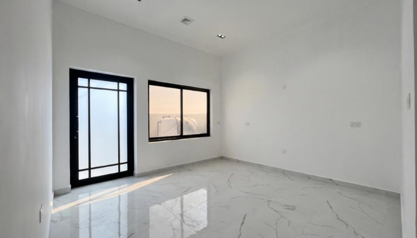 Empty room with white walls, large window, glass door, and marble floor tiles. Ceiling lights are on, and sunlight is coming through the window and door.