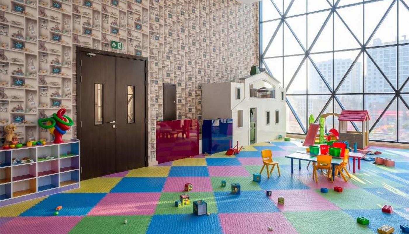 Spacious playroom with colorful foam floor tiles, scattered toys, playhouses, child-sized furniture, and large windows overlooking city buildings.
