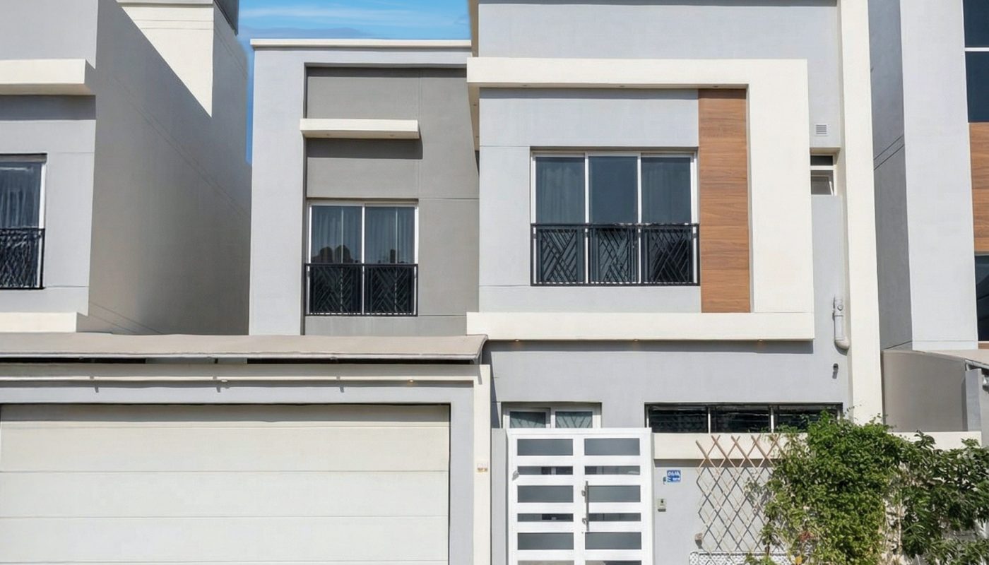 Modern two-story house with a flat roof, large garage door, white and grey exterior, and minimal landscaping in front under a clear blue sky.