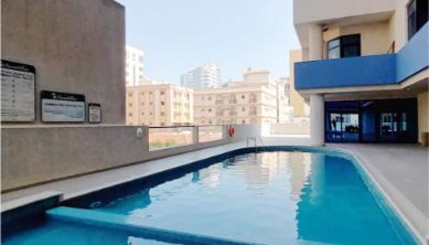 Outdoor rectangular swimming pool next to a multi-story apartment building, with safety signs and city buildings visible in the background.