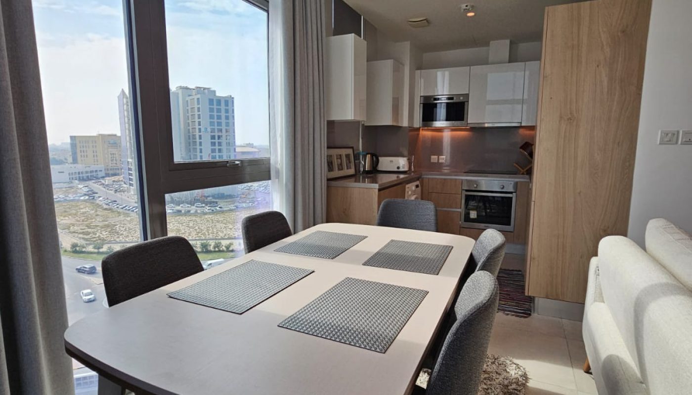 A modern apartment dining area with a table set for six, next to large windows, and an open kitchen with white cabinets and built-in appliances.