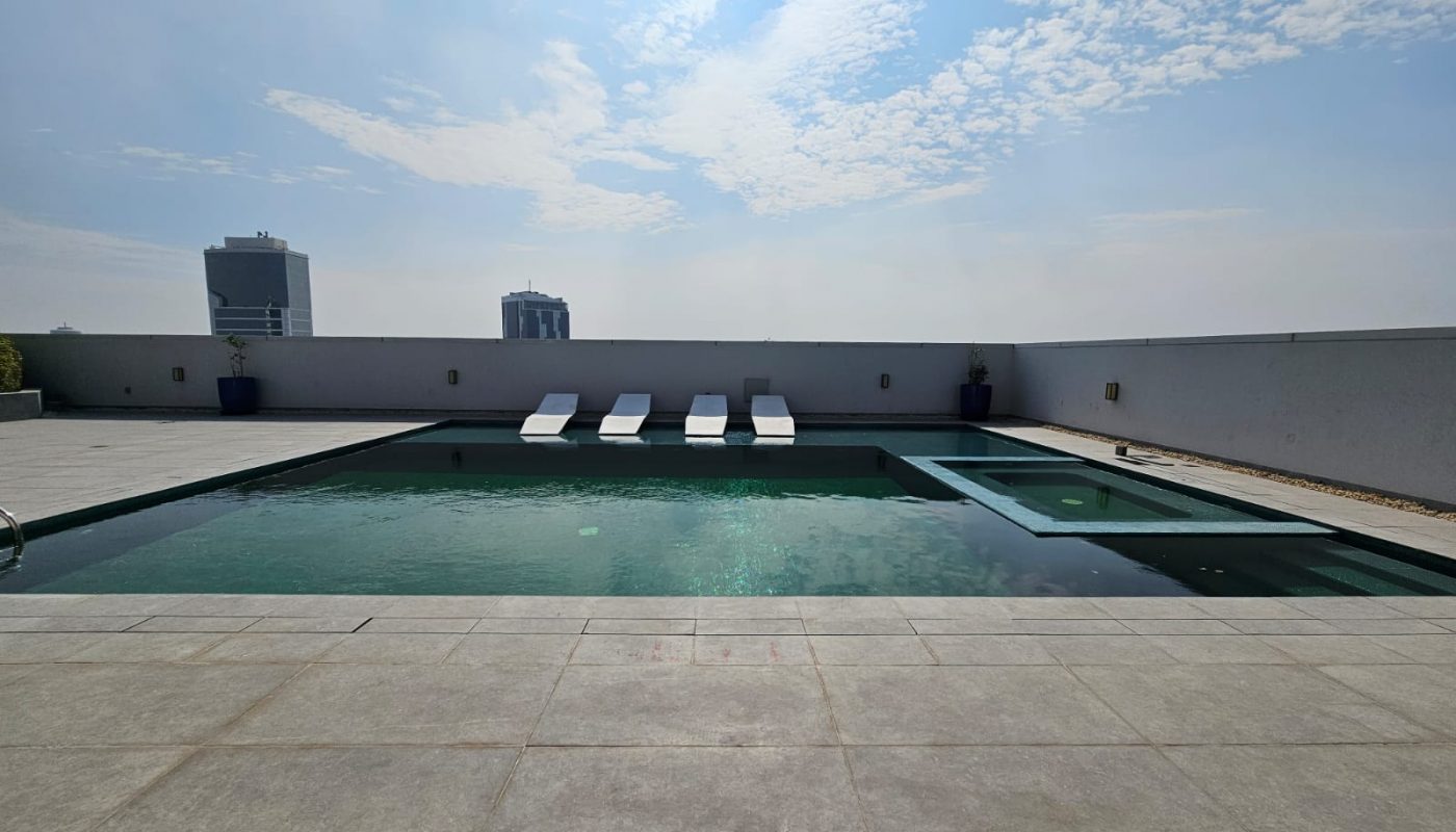 Outdoor pool with four white lounge chairs partially in the water, surrounded by a tiled deck, under a partly cloudy sky with city buildings in the background.
