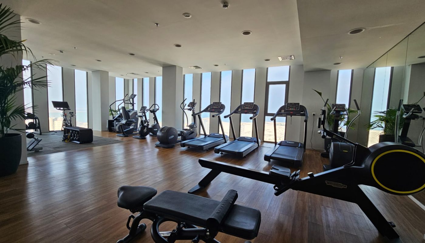 Modern gym with treadmills, rowing machine, stationary bikes, and strength equipment. Large windows let in natural light; wooden floors and mirrors enhance the spacious setting.