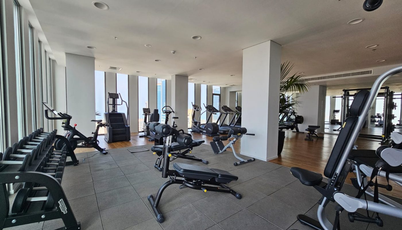 Modern gym with large windows, various exercise machines, free weights, and benches arranged on tiled and wooden flooring.
