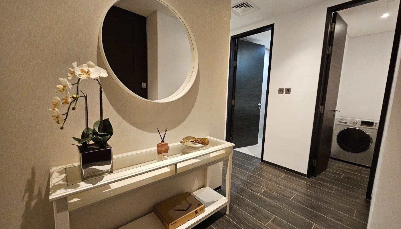 A fully-furnished hallway with a console table holding a plant, decorative items, and a mirror above. A door on the right leads to one of the 2 bedrooms in the flat, equipped with its own washing machine.
