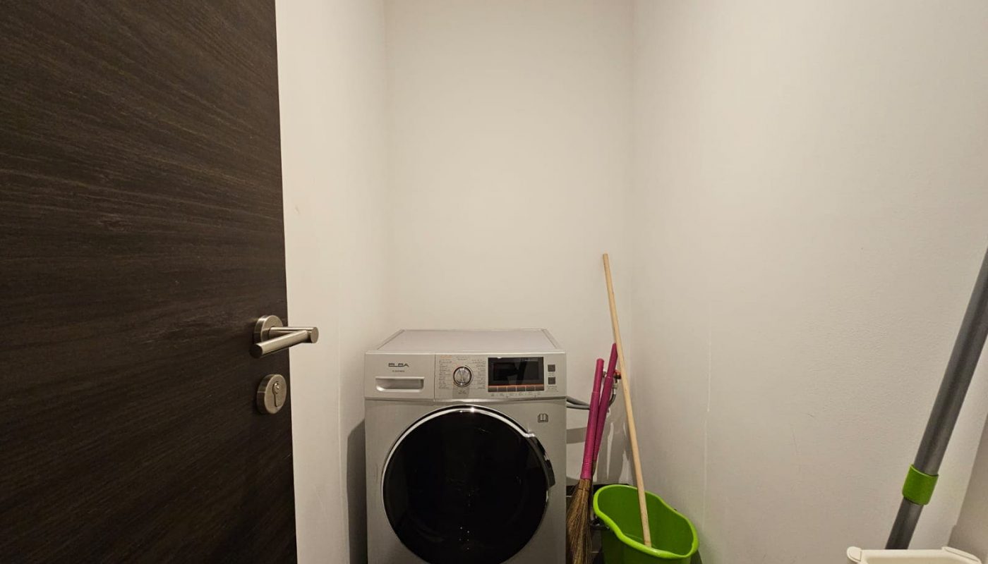A small laundry room in a fully-furnished flat features a washing machine, a mop, a green bucket, and a broom against a white wall. A closed dark wooden door to the spacious 2-bedroom is partially visible.