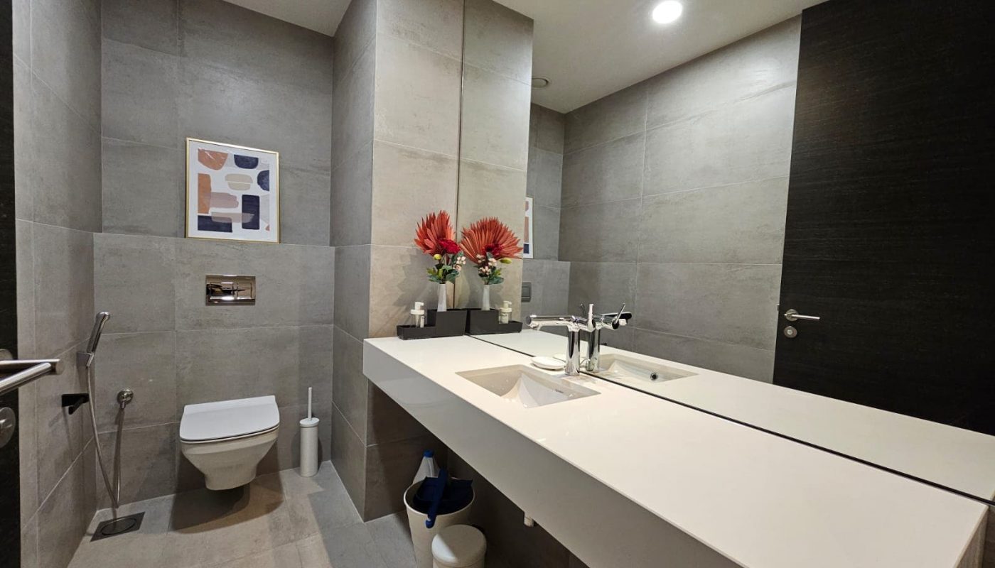 A modern bathroom in a spacious, fully-furnished flat, featuring a toilet, double sink vanity, mirror, and wall art. The decor includes red flowers on the sink and gray tiles on the walls and floor. This bathroom complements the stylish design of the 2-bedroom home.