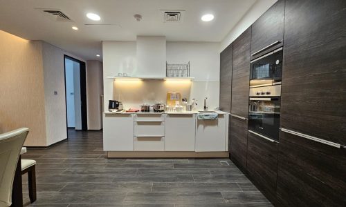 A modern kitchen in a spacious, fully-furnished flat with white cabinetry, dark floor tiles, and built-in appliances including an oven and microwave. The counter is equipped with a kettle, toaster, knife set, and various kitchen utensils.