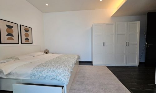 A minimalist fully-furnished bedroom with a double bed, white bedding, two abstract framed artworks on the wall, a white wardrobe, and a small table lamp on a nightstand. Perfect for any flat looking for modern elegance.