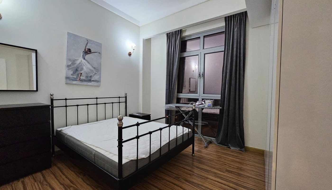 A neatly arranged bedroom in a flat for rent in Seef with a metal frame bed, wooden flooring, grey curtains, and a painting on the wall.