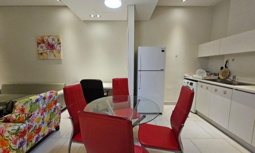 Modern kitchen with a glass dining table, red chairs, and white appliances available in a flat for rent in Seef.