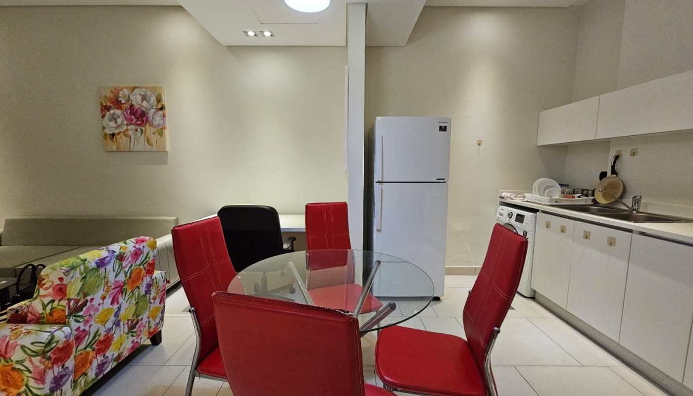 Modern kitchen with a glass dining table, red chairs, and white appliances available in a flat for rent in Seef.