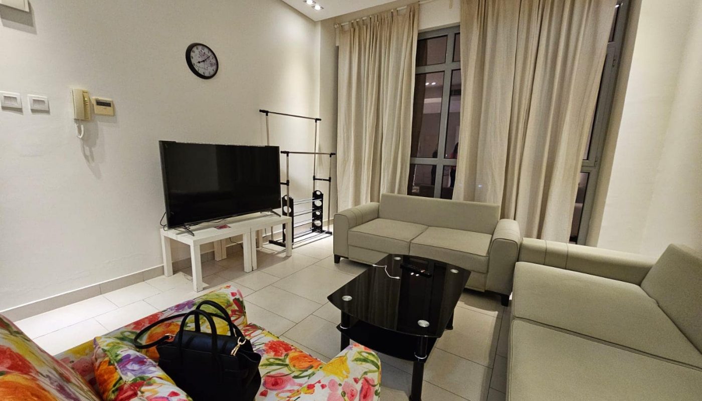 A modern living room interior at night with a flat-screen tv, a beige sofa, a colorful armchair, and glass center tables is available for rent in Seef.