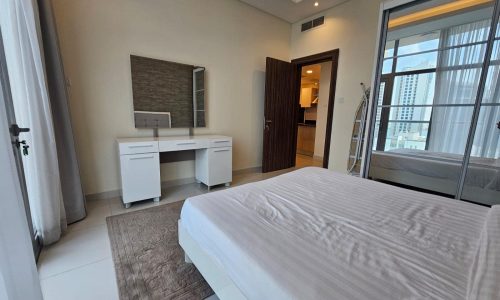 Modern bedroom in a luxury apartment with a neatly made bed, a desk, and sliding doors leading to a balcony in Juffair, for rent.