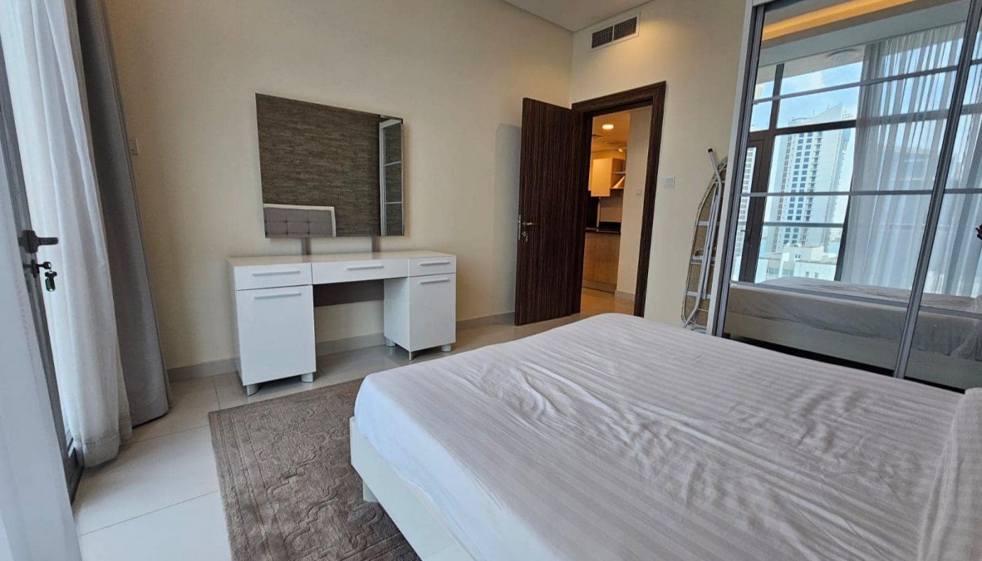 Modern bedroom in a luxury apartment with a neatly made bed, a desk, and sliding doors leading to a balcony in Juffair, for rent.