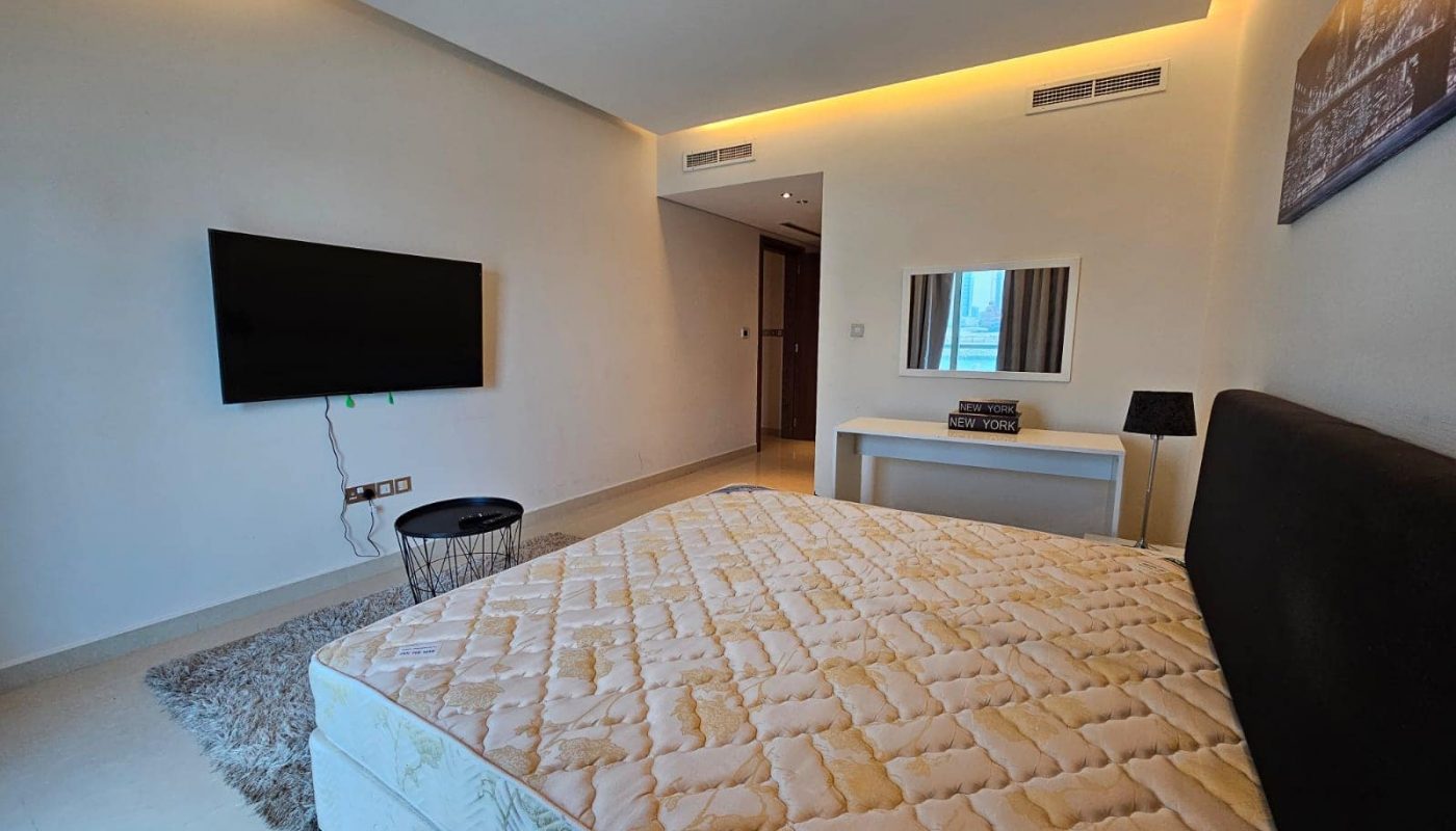 A modern, well-lit bedroom with a large bed, a wall-mounted television, and minimalistic decor.