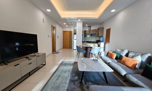 Luxurious modern living room with a gray sofa, television, and an open-plan kitchen in the background, available for rent in Juffair.