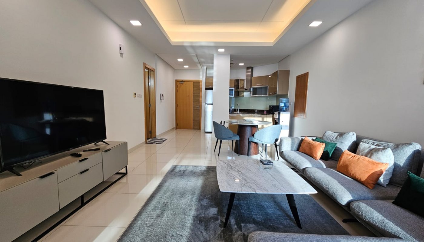 Luxurious modern living room with a gray sofa, television, and an open-plan kitchen in the background, available for rent in Juffair.
