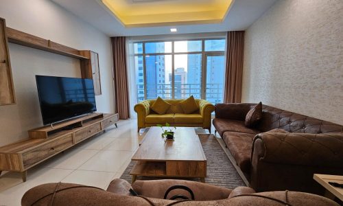 Modern living room interior with a brown sofa, yellow loveseat, and a television unit, overlooking a cityscape through the window. Auto Draft