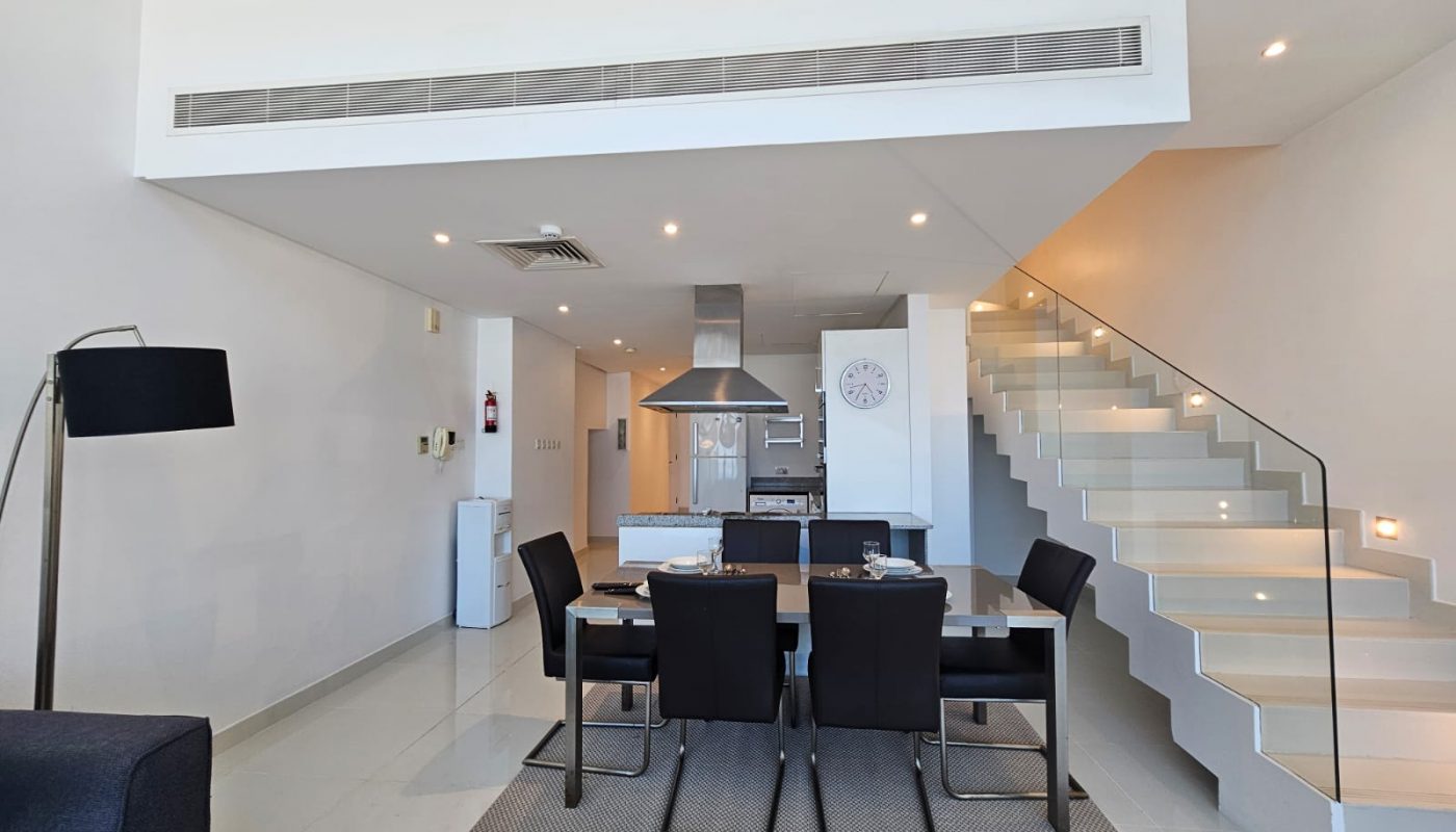 Modern dining area with black chairs and table, adjacent to a kitchen and staircase within a bright, minimalist interior.