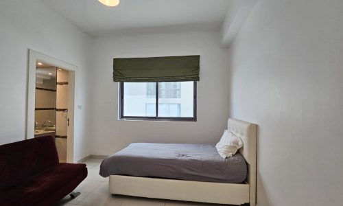 A modern concept bedroom within a fully-furnished 3-bedroom flat in Juffair, featuring a single bed, a red sofa, and a window with a green blind.