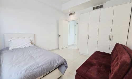 Minimalist bedroom in a spacious flat with a white bed, built-in wardrobes, and a red sofa.