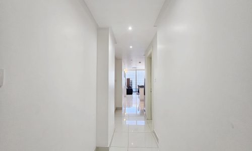 A bright, narrow white hallway with glossy tiles leading to a lit room at the end of a 3 Bedroom Fully-furnished Flat in Juffair.