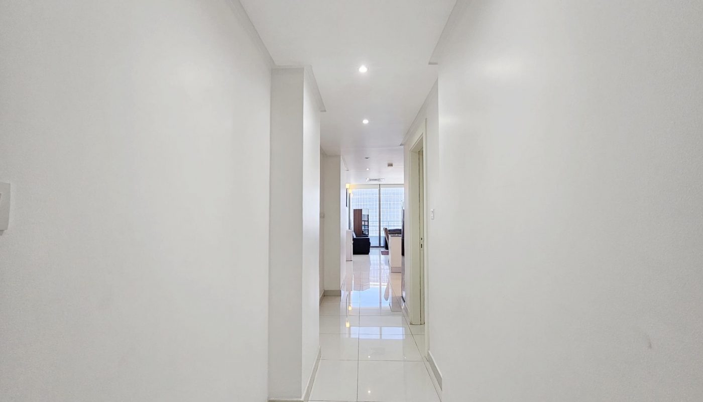 A bright, narrow white hallway with glossy tiles leading to a lit room at the end of a 3 Bedroom Fully-furnished Flat in Juffair.