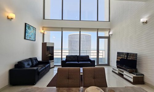 Modern living room interior in a fully-furnished flat located in Juffair, with large windows offering a city view, furnished with black sofas, a television, and a dining area.