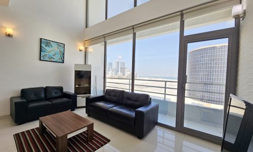 Modern living room in a 3-bedroom fully-furnished flat with black leather sofas, a wooden coffee table, and a large window offering a city view.