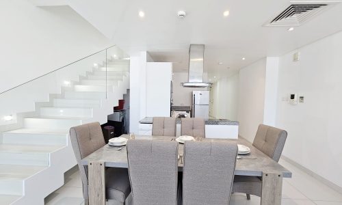 Modern dining room in a fully-furnished, 3-bedroom flat located in Juffair, with a table set for four, leading to a kitchen and staircase in a bright interior.