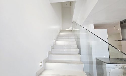 A clean, modern concept interior with a white staircase and glass balustrade, leading up to a well-lit, fully-furnished flat in Juffair.