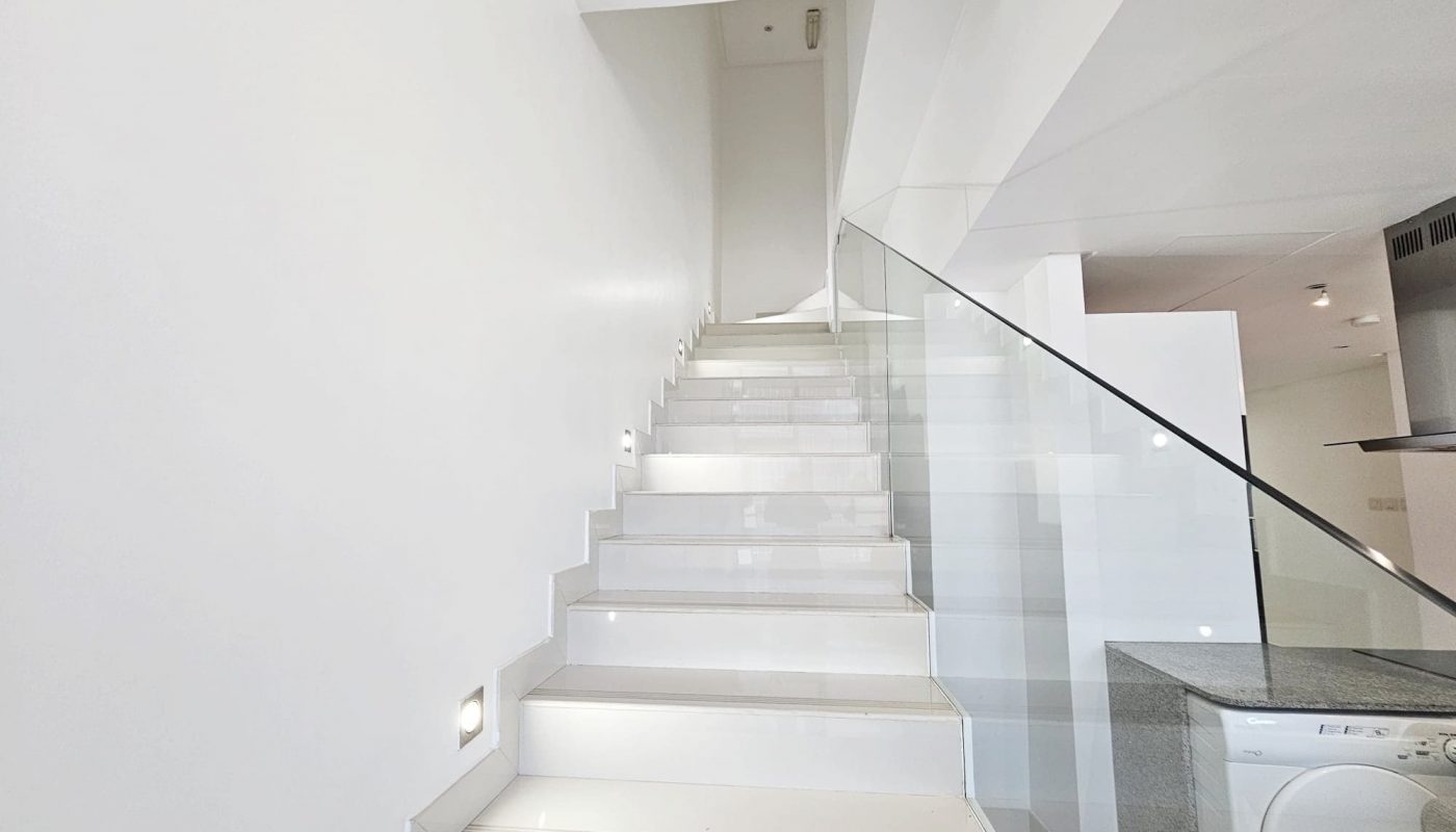 A clean, modern concept interior with a white staircase and glass balustrade, leading up to a well-lit, fully-furnished flat in Juffair.