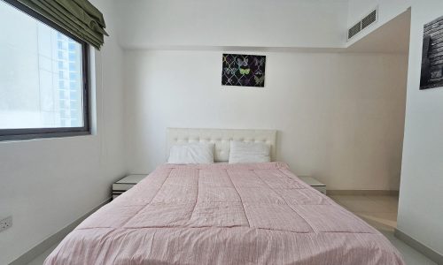 A neatly arranged bedroom in a 3 Bedroom Fully-furnished Flat in Juffair with a large bed covered with a pink bedspread, simple decor, and a window allowing natural light, designed according