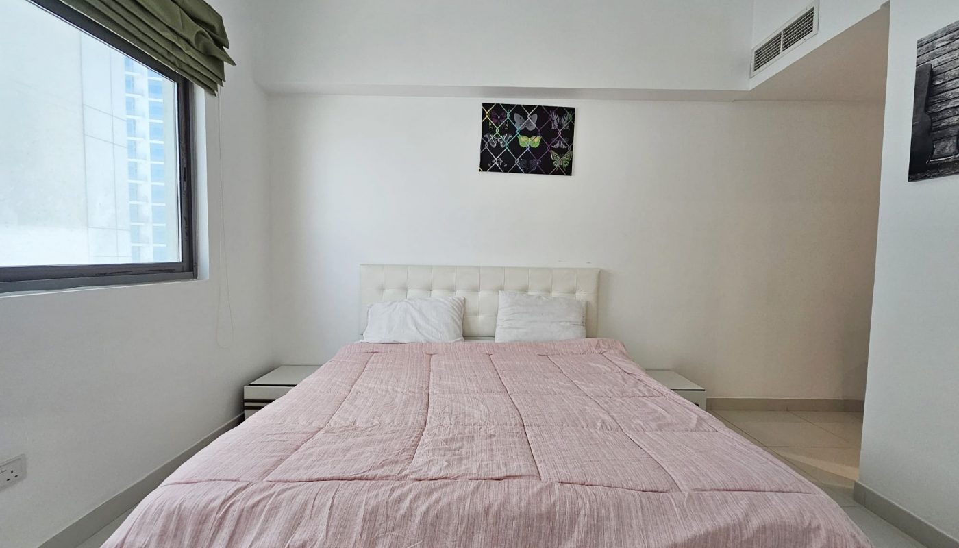 A neatly arranged bedroom in a 3 Bedroom Fully-furnished Flat in Juffair with a large bed covered with a pink bedspread, simple decor, and a window allowing natural light, designed according