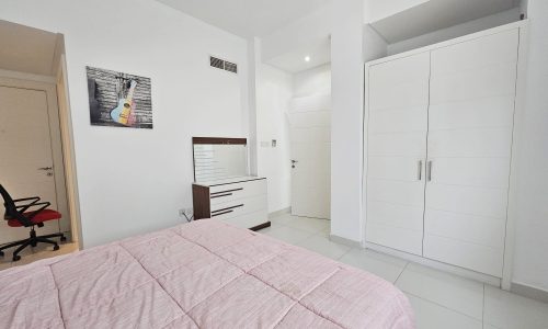 A fully-furnished bedroom with a bed covered in a pink bedspread, white furniture, and a wall-mounted picture.