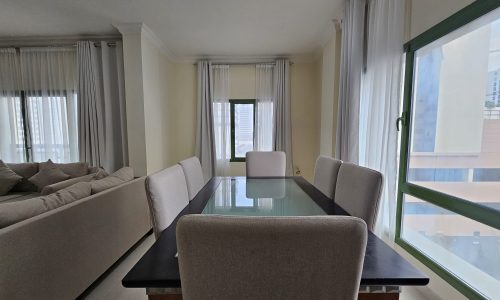 Modern 2BR apartment interior in Juffair featuring a dining table set near a balcony with sheer curtains and a peek of urban views.