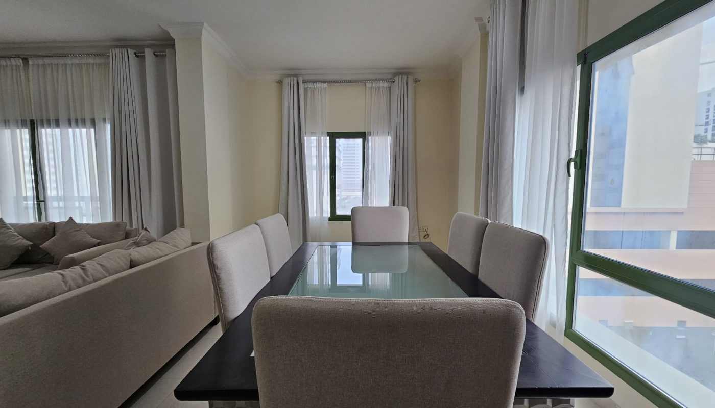 Modern 2BR apartment interior in Juffair featuring a dining table set near a balcony with sheer curtains and a peek of urban views.