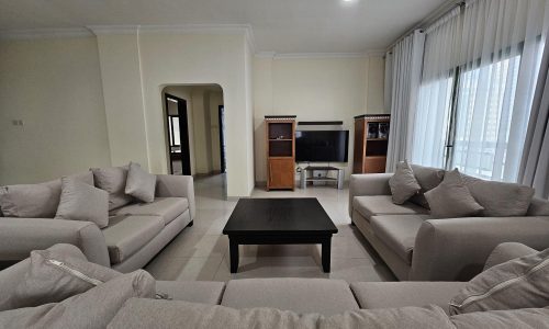 A modern 2BR apartment in Juffair with a living room featuring multiple beige sofas and a central black coffee table, with a television set against the wall available for rent.