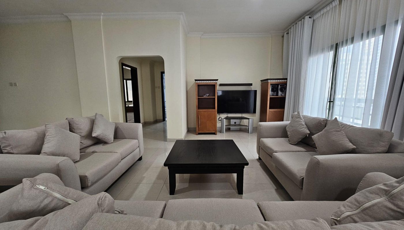 A modern 2BR apartment in Juffair with a living room featuring multiple beige sofas and a central black coffee table, with a television set against the wall available for rent.